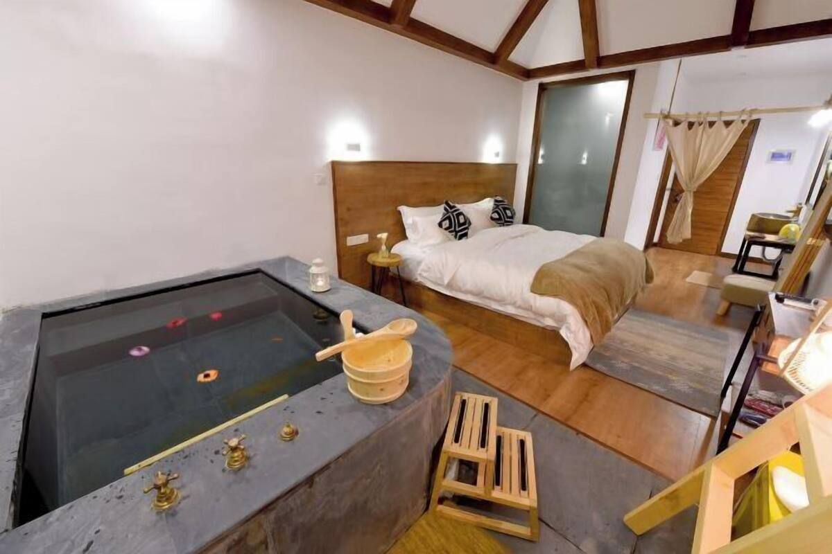 Un dormitorio con una cama grande, cabecero de madera, un taburete pequeño de madera y una bañera de piedra con lavabo de madera.