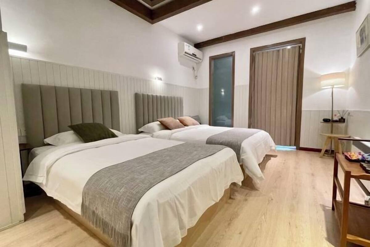Habitación de hotel con dos camas, suelo de madera, una ventana con cortinas, un equipo de aire acondicionado y una mesita con lámpara.