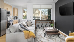 Un salon moderne avec un canapé, une table basse et un téléviseur fixé au mur. Il y a également un coin cuisine équipé d’un réfrigérateur, d’un four à micro-ondes et de placards. La pièce est dotée de grandes fenêtres et offre une vue sur une maison à l’extérieur.