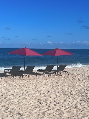 A beach with three lounge chairs and two red umbrellas.