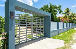 A gated entrance with a white fence, a concrete pathway, and lush greenery in the background.