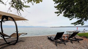 Uma área de praia com pedrinhas, com duas cadeiras de descanso e uma mesa sob um toldo, com vista para um lago calmo e montanhas ao longe.
