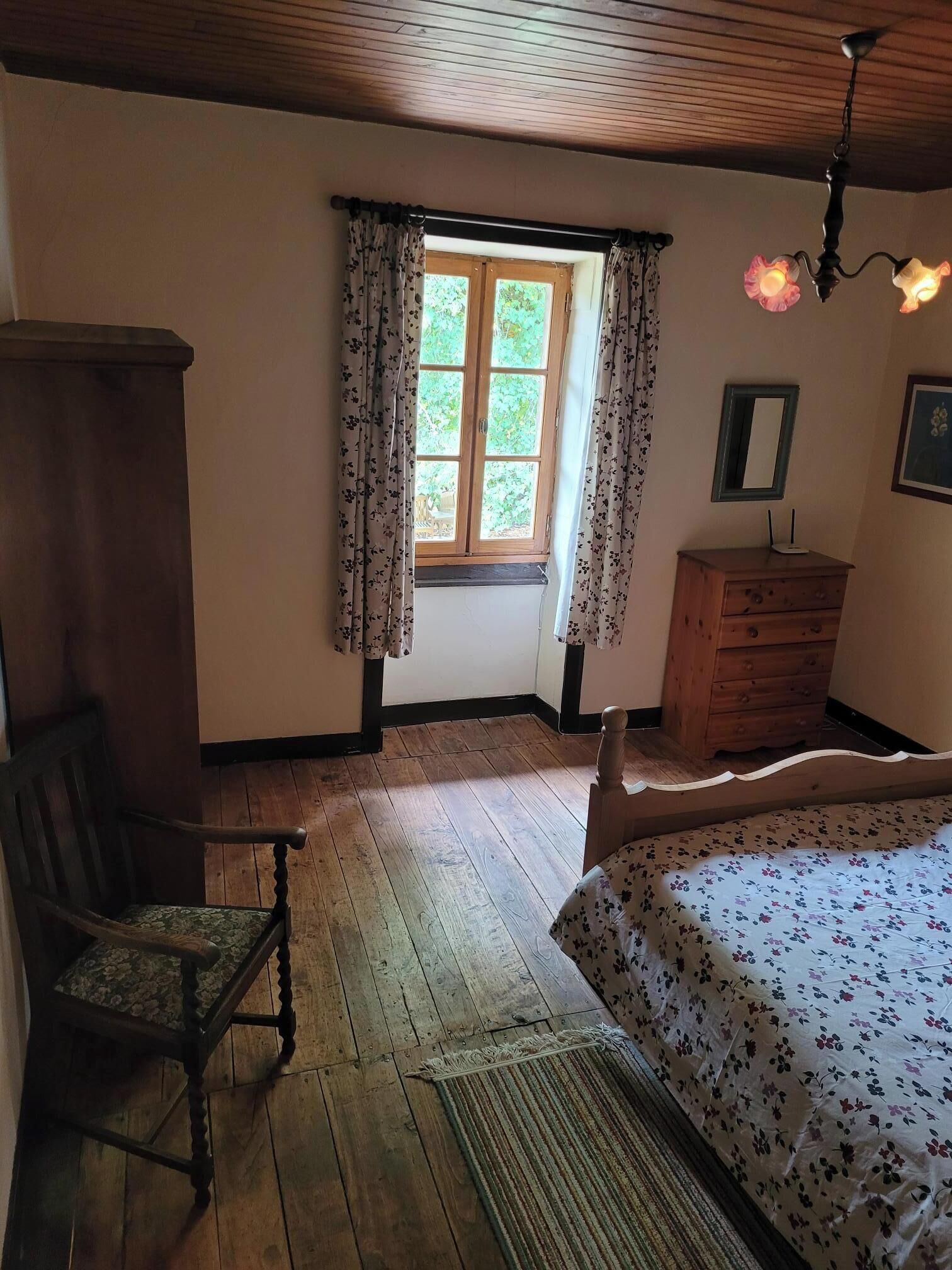 A bedroom with a wooden floor, a bed with floral bedding, a wooden dresser, a chair, a window with floral curtains, and a ceiling light fixture.