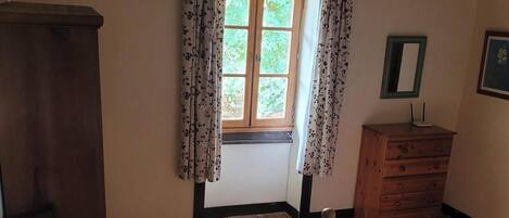 A bedroom with a wooden floor, a bed with floral bedding, a wooden dresser, a chair, a window with floral curtains, and a ceiling light fixture.