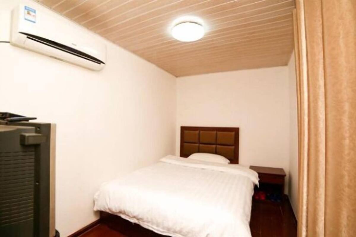 A small, clean room with a bed, a nightstand, a wooden ceiling, and an air conditioning unit.