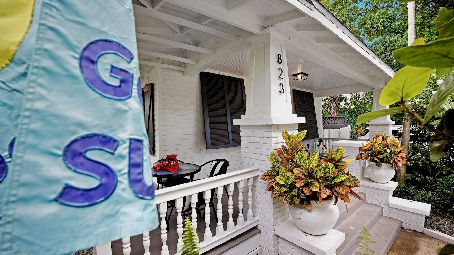 A white porch with a number 823 sign, a table with chairs, and potted plants.