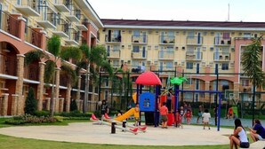 A playground with slides and swings, surrounded by a building with balconies and a palm tree. - Lian's Haven at Arezzo Place Davao (Davao City)