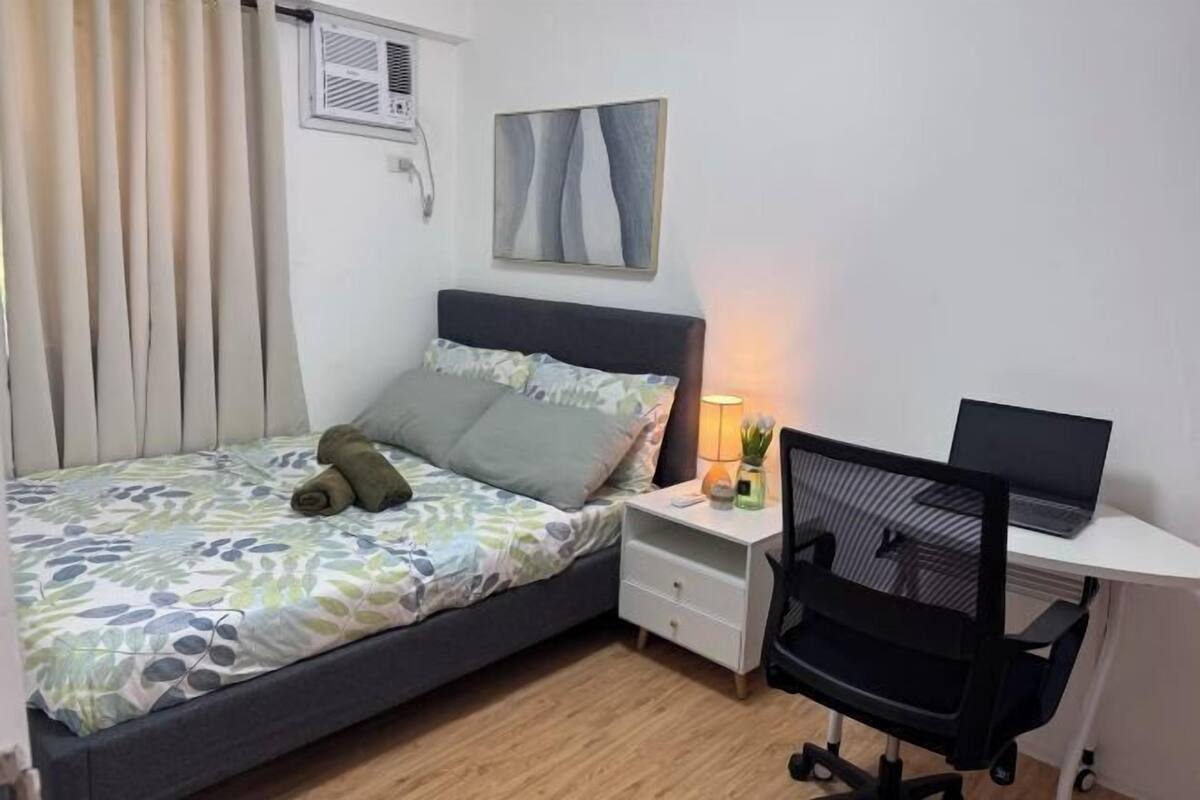 Une chambre à coucher comprenant un lit, un bureau avec un ordinateur portable, une chaise, une table de chevet et un climatiseur fixé au mur.