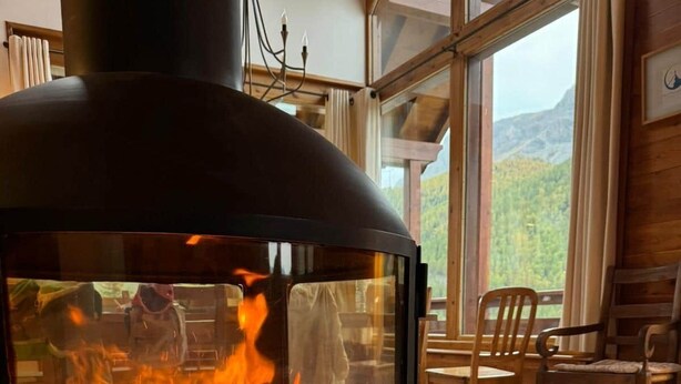 A wood-burning stove with a fire, a view of the outdoors, and a dining area with chairs and a table. - Chalet face aux étoiles (Molines-en-Queyras)