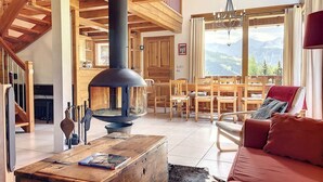 A cozy living room with a wooden ceiling, a large fireplace, a dining area with wooden furniture, and a view of mountains through large windows. - Chalet face aux étoiles (Molines-en-Queyras)