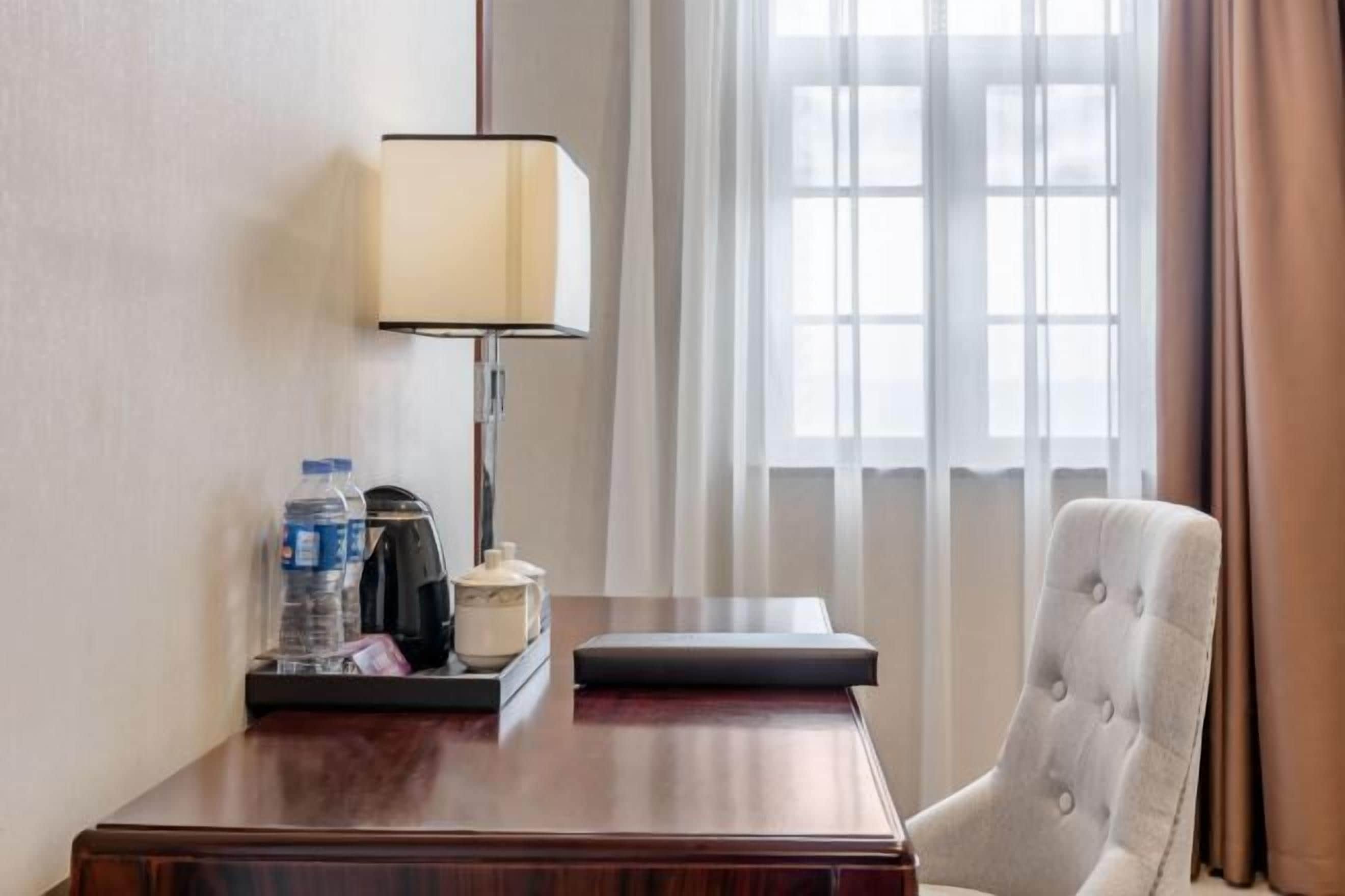 A hotel room with a wooden desk, a lamp, a chair, a window with curtains, and a view of the outside.