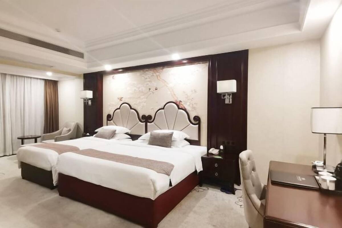 A hotel room with a large bed, two bedside tables, a desk with a chair, a chair, and a wall-mounted lamp.