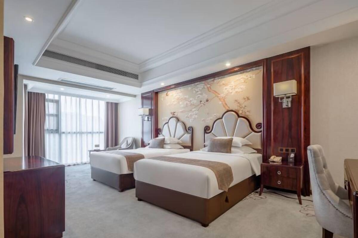 A hotel room with two beds, a large window, a decorative wall, and a desk with a lamp.