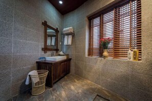 A bathroom with a stone wall, wooden vanity, a hanging mirror, a window with blinds, and a basket. - Yunmengze Hostel (Lijiang)