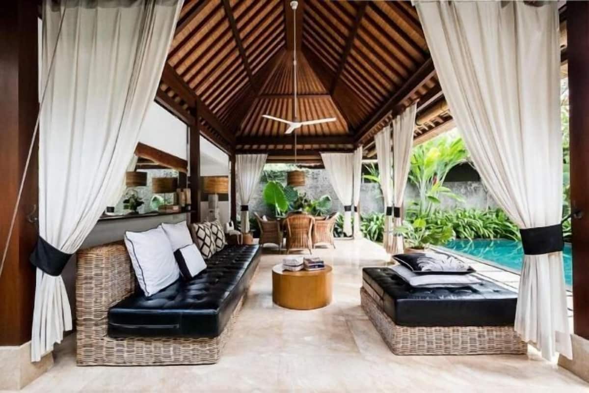 A covered outdoor seating area with wicker furniture, a central round table, and a view of a pool and greenery.