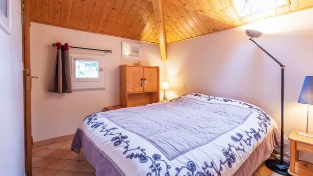 Ein Schlafzimmer mit Holzboden, einem Bett mit gemustertem Überwurf, einem Nachttisch mit Lampe, einem Holzschrank und einem Fenster mit Vorhängen.