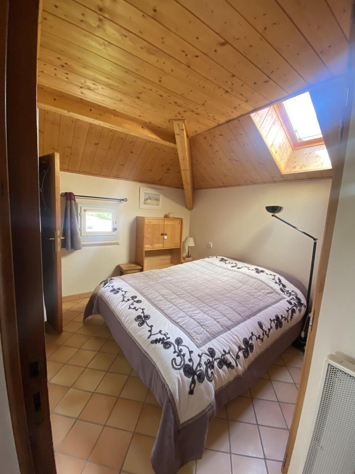 A bedroom with a sloped ceiling, a bed with a floral bedspread, a small wooden cabinet, a nightstand, a lamp, and a window with a view of the outside.
