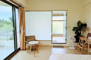 Habitación de hotel con cama, una mesita, una silla, un ventilador y un ventanal con cortinas.