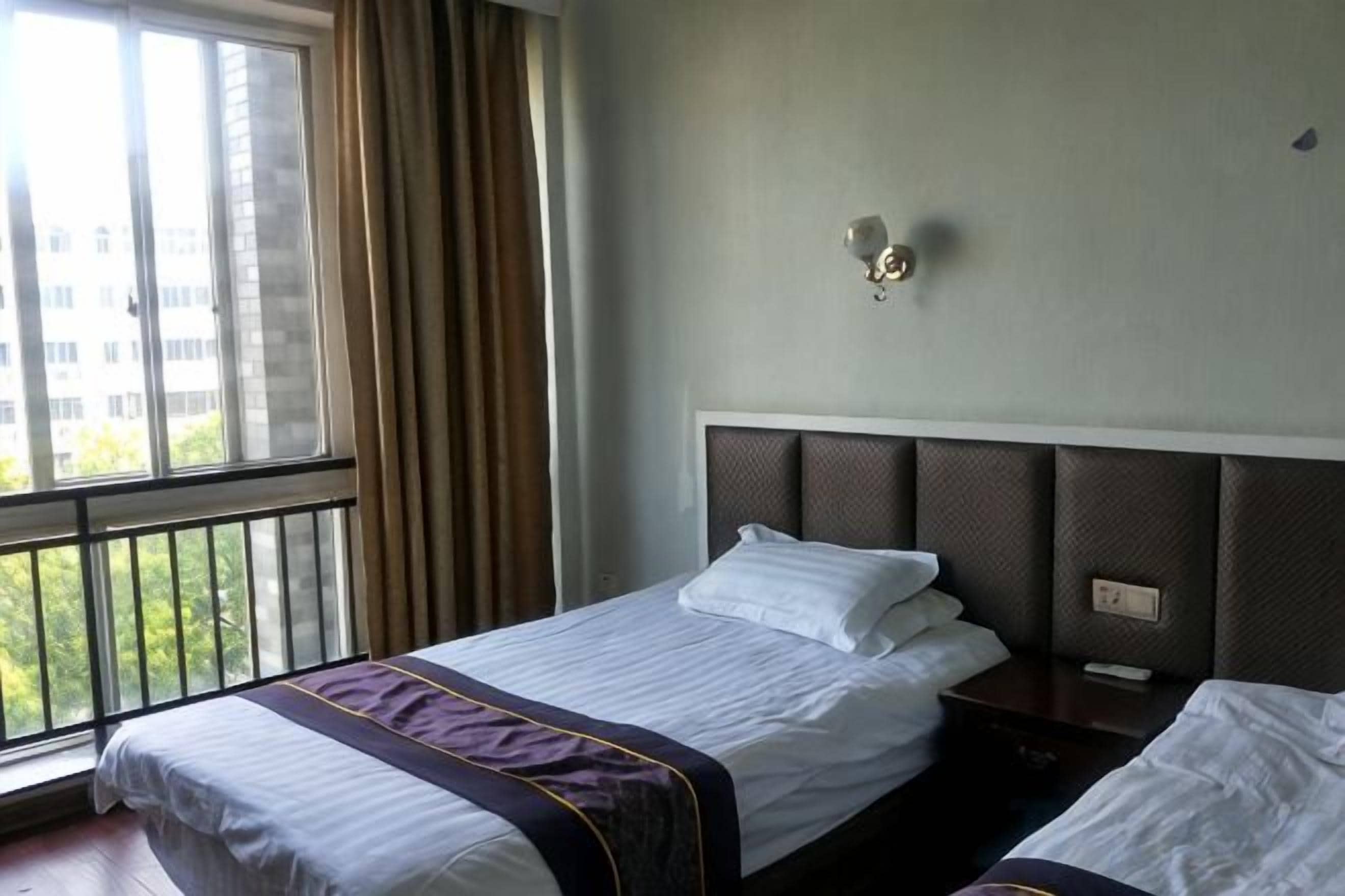 Habitación de hotel con cama, mesita de noche, ventana con cortinas y vistas a edificios.