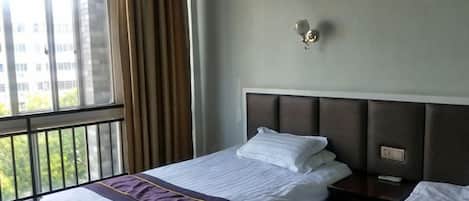 A hotel room with a bed, a nightstand, a window with curtains, and a view of buildings outside.