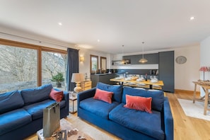 A modern living room with a large blue sofa, a dining area with a wooden table, and a kitchen in the background.