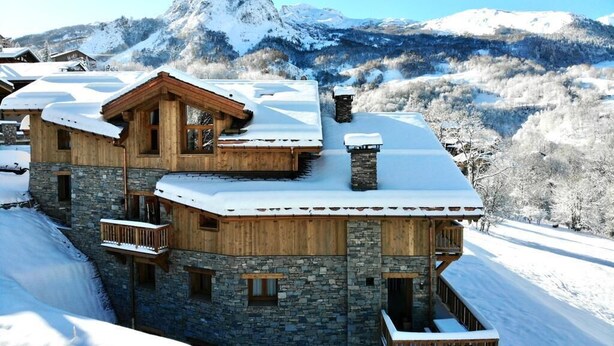 Un chalet in legno a più piani con pareti in pietra, tetti imbiancati dalla neve e balconi, sullo sfondo di montagne.