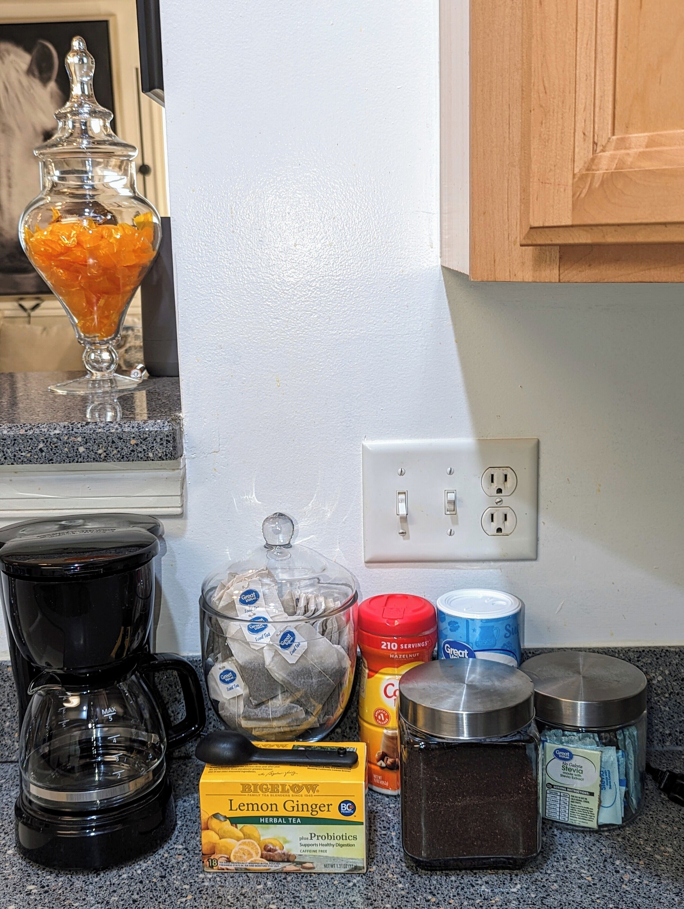 A kitchen counter with a coffee maker, a glass jar with orange items, a box of lemon ginger herbal tea, a container of hazelnut coffee, and a can of sweet tea.