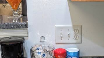 A kitchen counter with a coffee maker, a glass jar with orange items, a box of lemon ginger herbal tea, a container of hazelnut coffee, and a can of sweet tea.