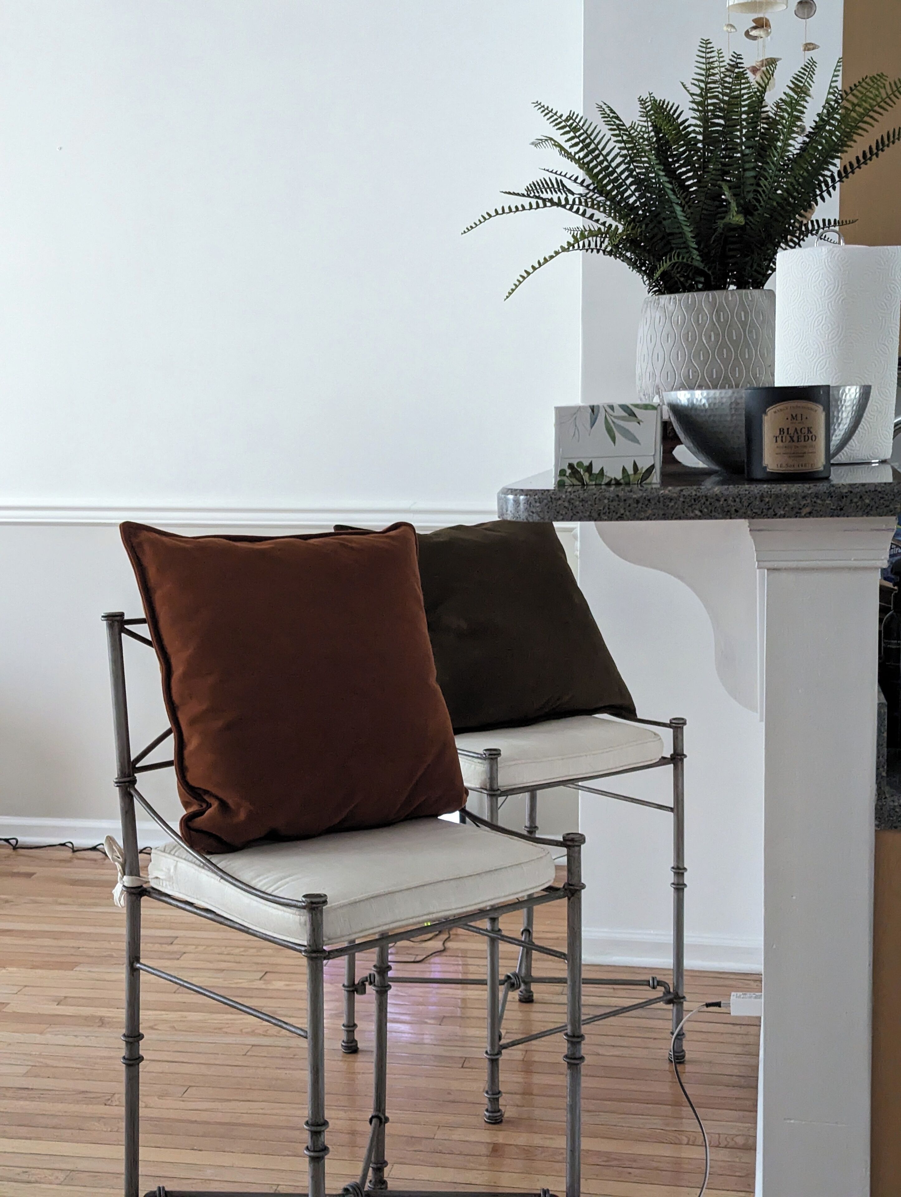A chair with a white cushion and a brown cushion, a potted fern, and a container with a label on a kitchen counter.