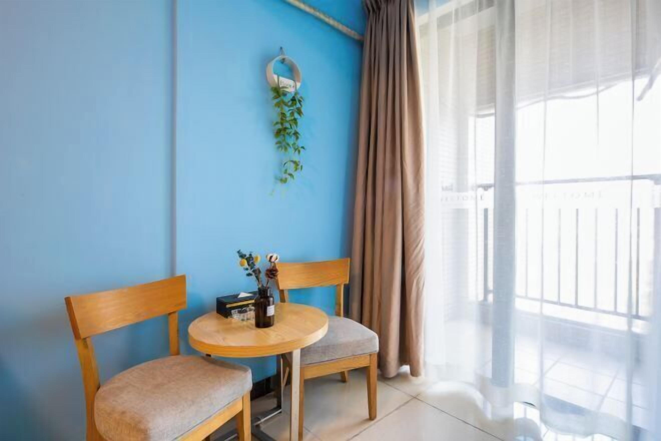 A room with a blue wall, a round wooden table with two chairs, a hanging plant, and a view through sheer curtains.