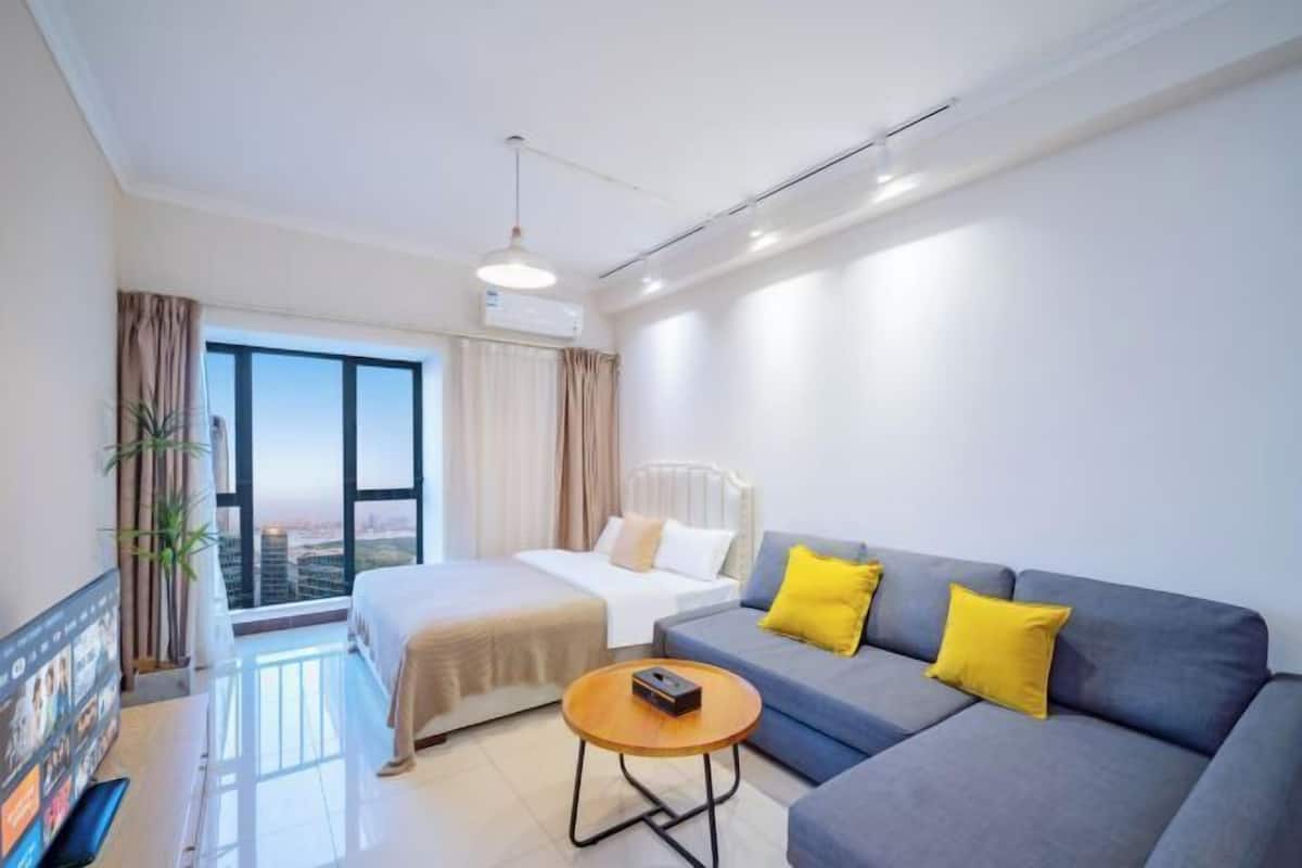 Quarto de hotel com cama, sofá, mesa de centro e uma grande janela com cortinas.