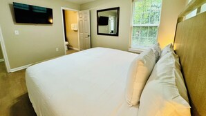 A bedroom with a large bed, a ceiling fan, a TV mounted on the wall, a window with blinds, and a door leading to a bathroom. - Modern Brick Ranch Retreat NEW Renovation (Mableton)