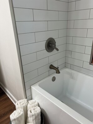 A bathroom with a white bathtub, a shower with a wall-mounted faucet, and a towel rack with white towels. - Whatchamacallit Haus, Downtown Shiner House (Shiner)