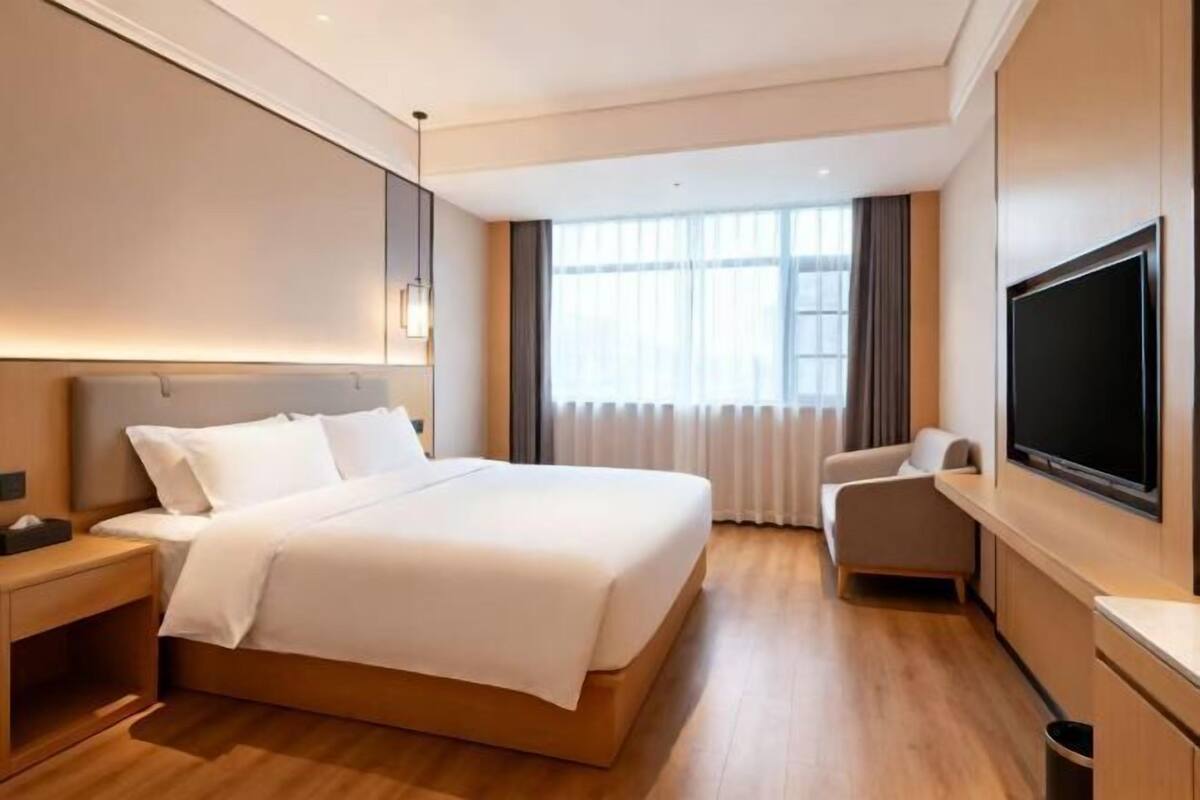 A modern hotel room with a large bed, a TV, a desk, and a window with curtains.