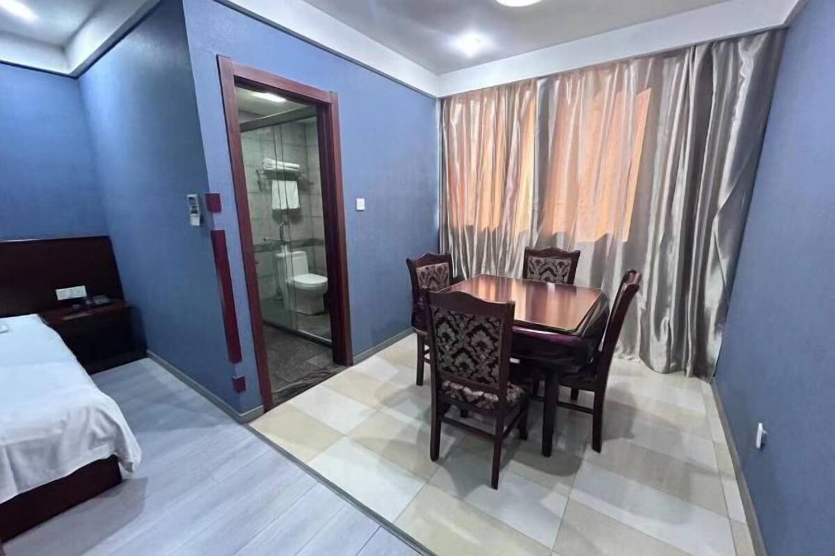 A hotel room with a bed, a dining table with chairs, a bathroom visible through a glass door, and curtains on the window.