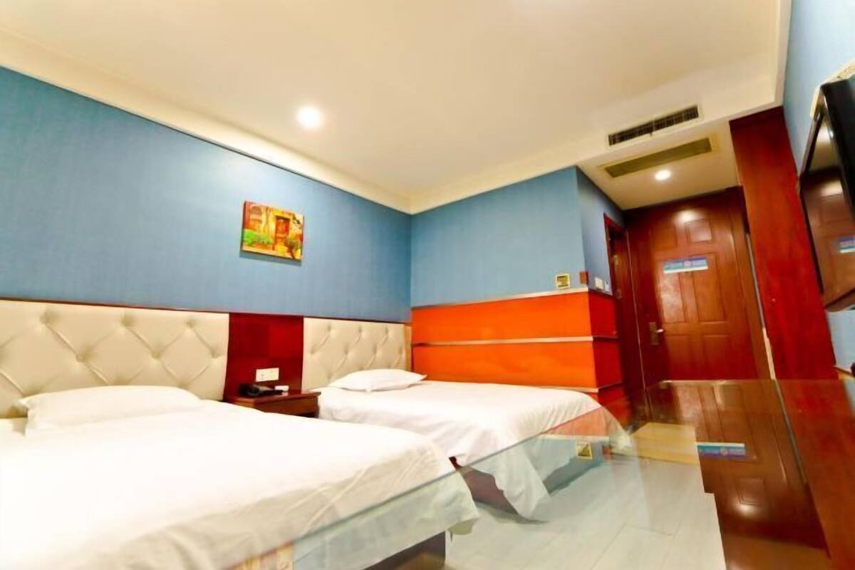 A hotel room with two beds, a glass partition, a red accent wall, and a wooden door.