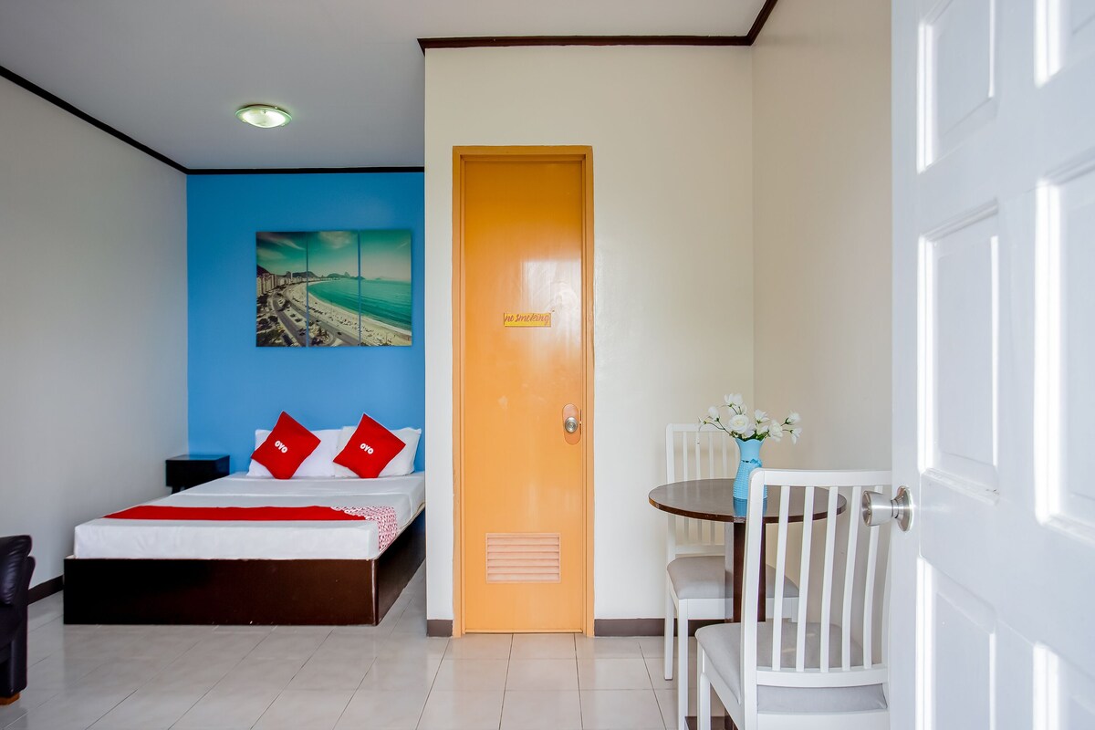 Quarto de hotel com uma cama, uma pequena mesa com cadeiras, um quadro na parede e uma porta com placa de 'proibido fumar'.