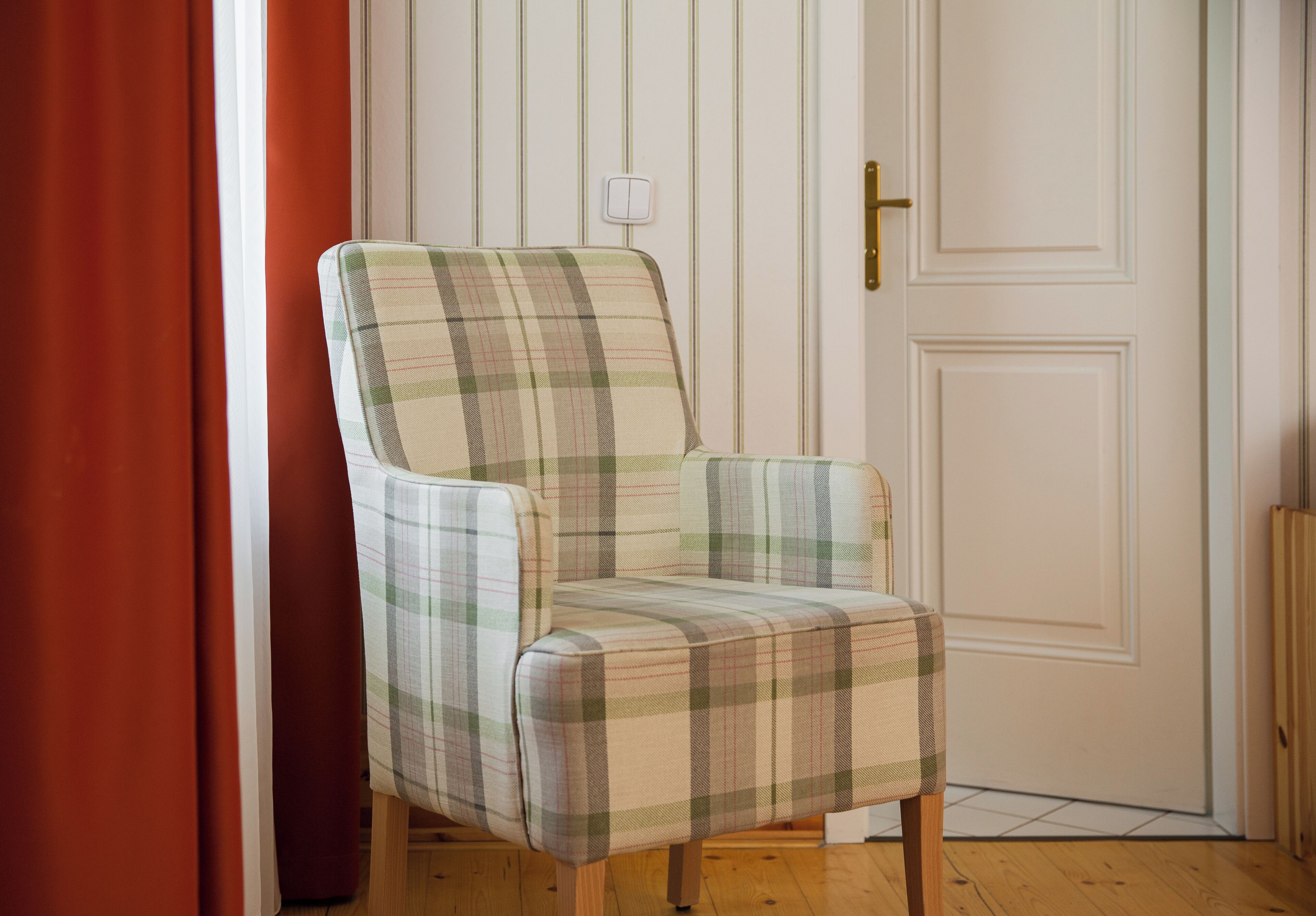A plaid-patterned armchair in a room with wooden flooring, a white door, and a red curtain.