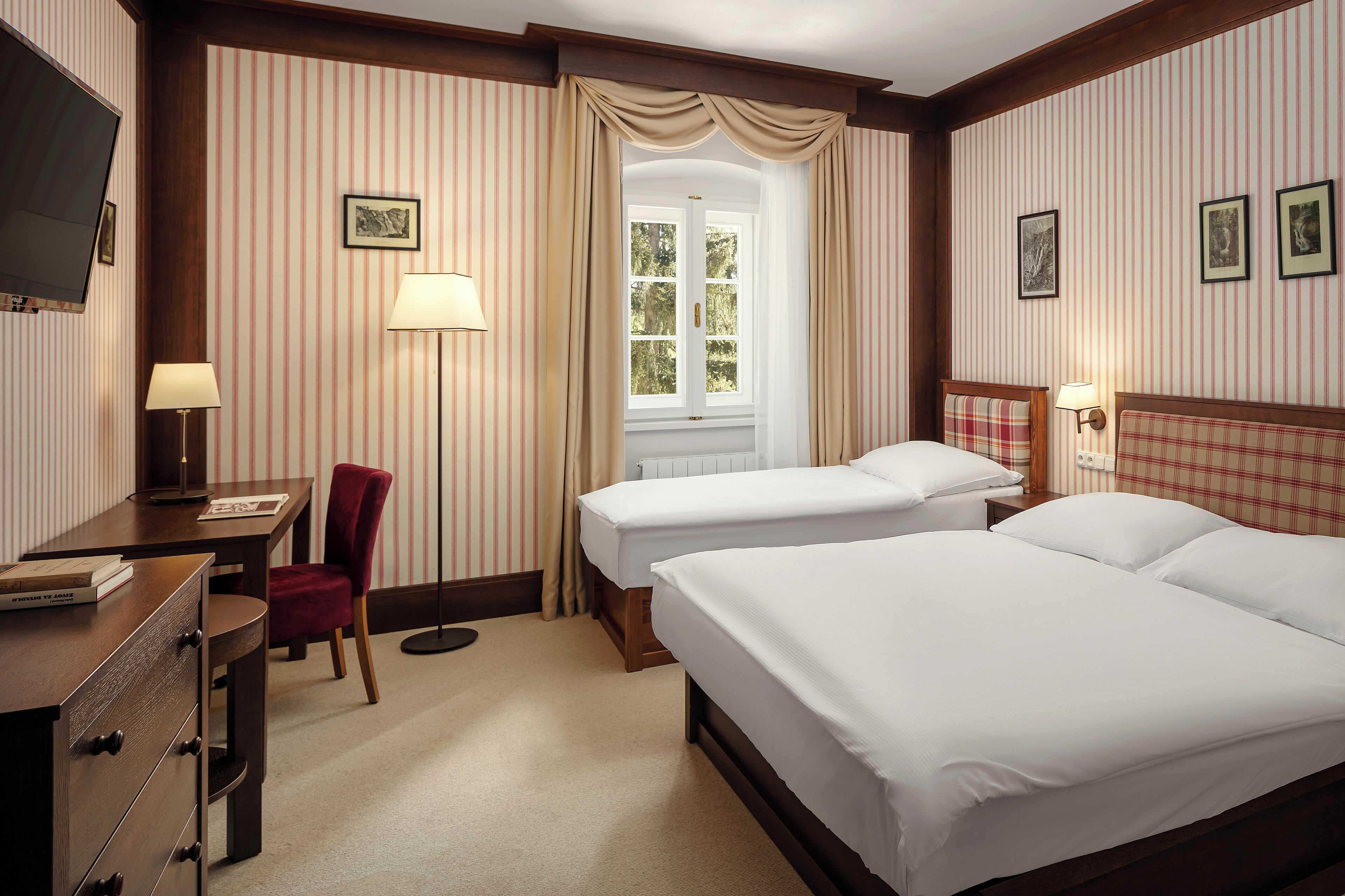 A hotel room with two beds, a desk, a chair, a TV, and a window with curtains.
