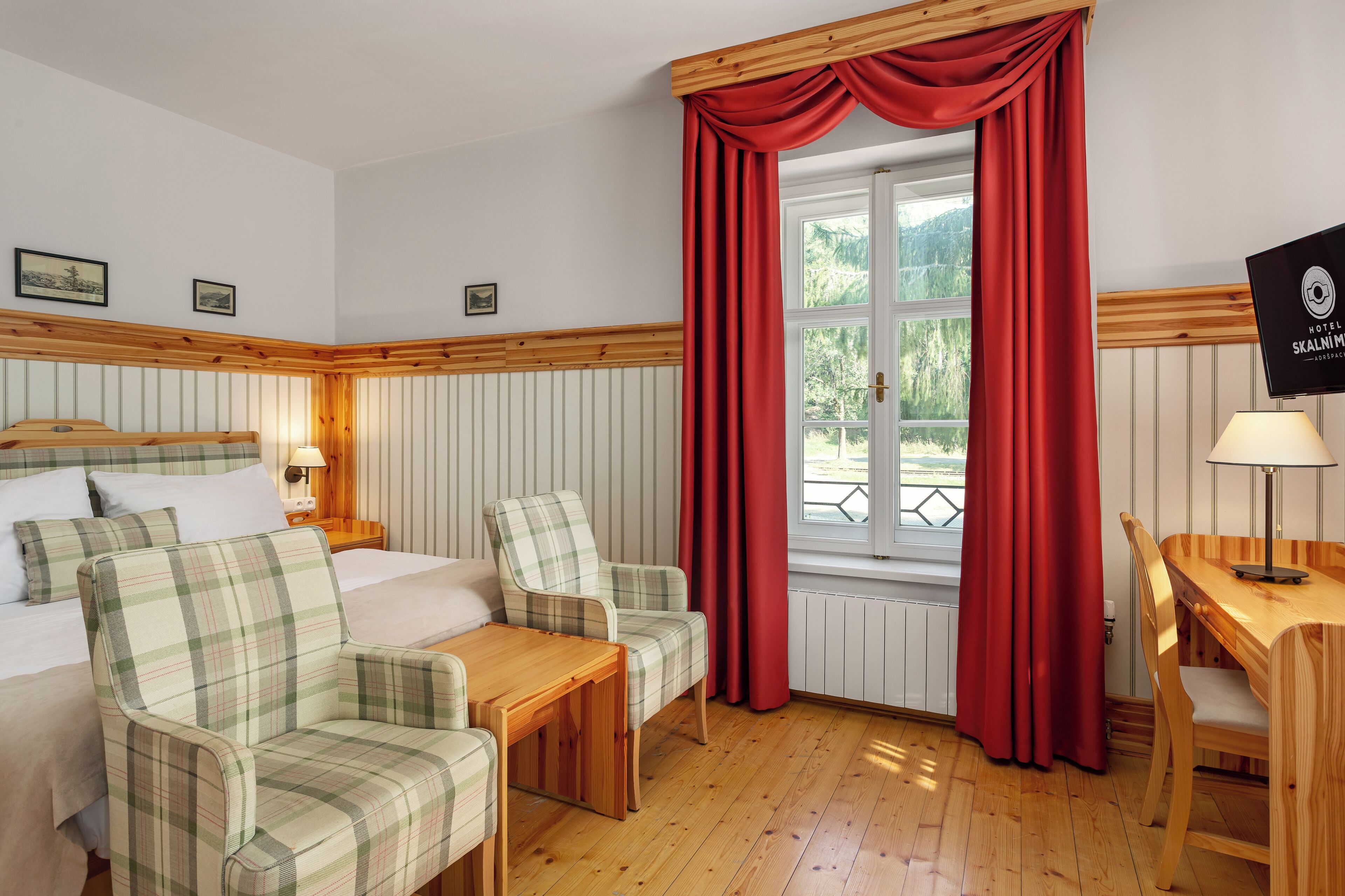 A hotel room with a bed, two plaid chairs, a wooden table, a window with red curtains, and a TV on the wall.