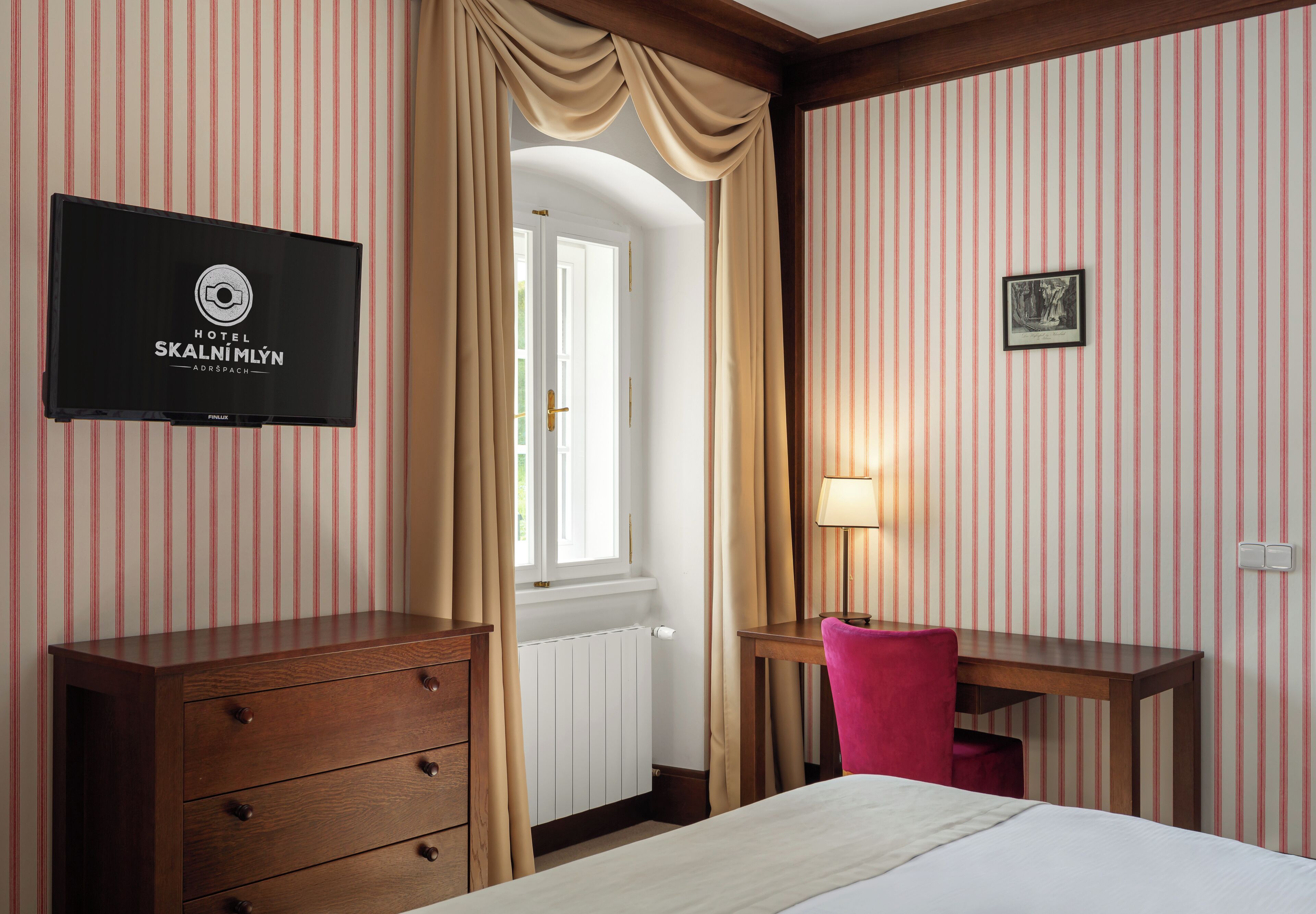 Habitación de hotel con papel tapiz a rayas, un aparador de madera, un escritorio con una silla, una cama, un televisor montado en la pared y una ventana con cortinas.