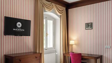 Habitación de hotel con papel tapiz a rayas, un aparador de madera, un escritorio con una silla, una cama, un televisor montado en la pared y una ventana con cortinas.
