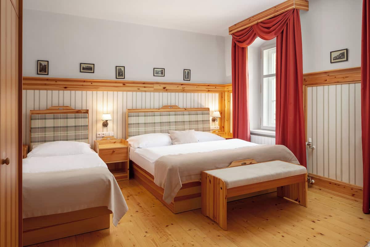 Habitación de hotel con dos camas, cabeceros de madera, un banco y una ventana con cortinas rojas.