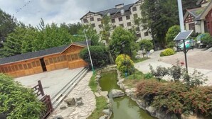 A traditional wooden building with a stone pathway leading to a small pond, surrounded by greenery and modern buildings in the background. - Lichuan Chunhuiyuan Homestay (Lichuan)