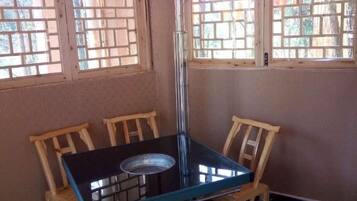 Una habitación con una mesa de vidrio y dos sillas de madera, ventanas amplias con marcos de madera y un radiador en el suelo.