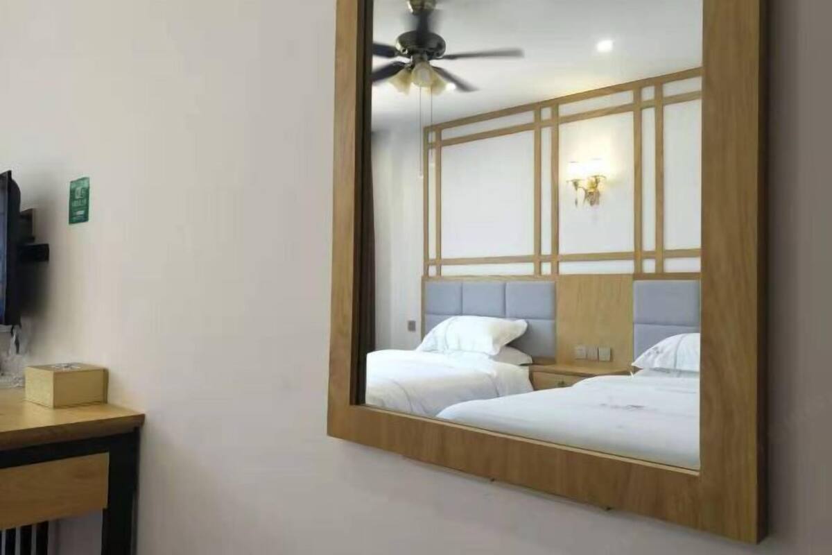 Habitación de hotel con un espejo grande que refleja dos camas, un ventilador de techo, una mesita de noche con lámpara y un televisor montado en la pared.