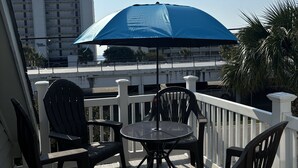 A balcony with two chairs and a table, a blue umbrella, and a view of a building with balconies. - Charming 3-bedroom house in North Myrtle Beach with AC, WiFi, pool and hot tub (North Myrtle Beach)