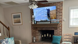 A living room with a brick fireplace, a television mounted above it, a ceiling fan, and a staircase leading to another level. - Charming 3-bedroom house in North Myrtle Beach with AC, WiFi, pool and hot tub (North Myrtle Beach)
