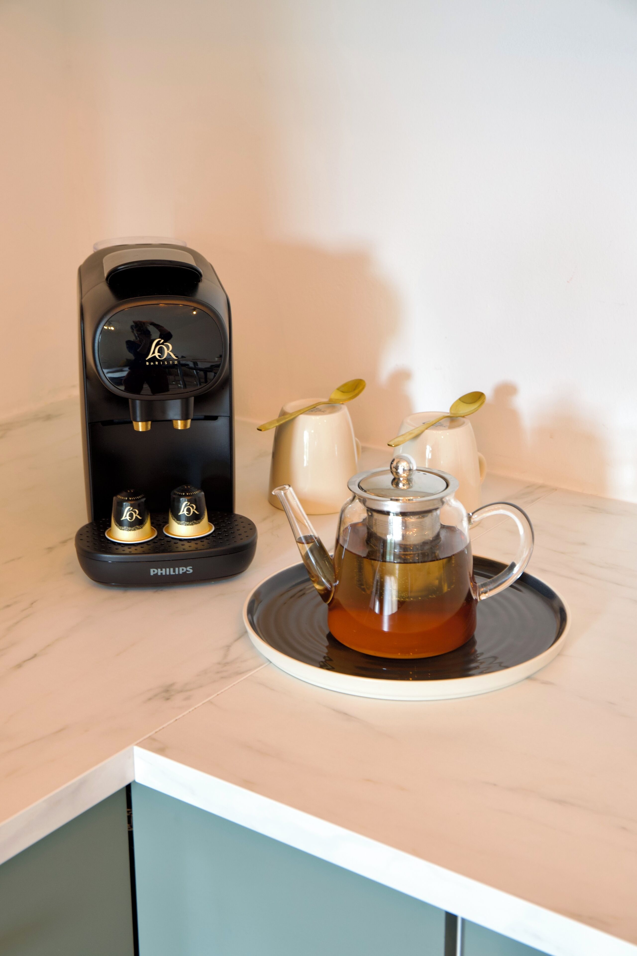 Une machine à café, une théière en verre et deux tasses blanches sur un plan de travail en marbre.