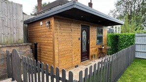 A small wooden cabin with a dark roof, a door with a glass panel, and a small patio area with a table and chairs.